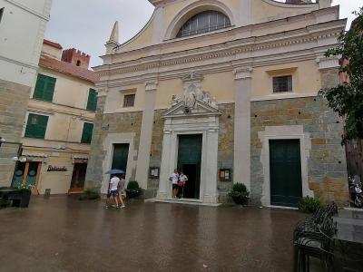 The image shows the facade of a church with a stone lower section and a plastered upper section, featuring a central pedimented doorway with statues above it, set within a paved square with people walking under an umbrella; a sign for "Ristorante"