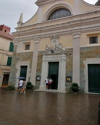 The image shows the facade of a church with a stone lower section and a plastered upper section, featuring a central pedimented doorway with statues above it, set within a paved square with people walking under an umbrella; a sign for "Ristorante"