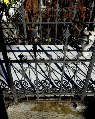 A close-up view of a white marble tombstone behind a black wrought-iron fence, with inscriptions for "GIUSEPPE TOMASI PRINCE DI LAMPEDUSA" and "ALESSANDRA WOLFF STEMMER PRINCESSA DI LAMPEDUSA".