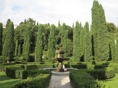A formal garden scene featuring a central stone fountain surrounded by low, geometrically trimmed boxwood hedges and tall, slender cypress trees under a partly cloudy sky.