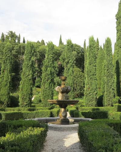 A formal garden scene featuring a central stone fountain surrounded by low, geometrically trimmed boxwood hedges and tall, slender cypress trees under a partly cloudy sky.