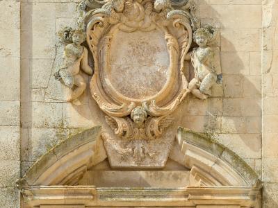 A close-up photograph of an ornate Baroque stone coat of arms and doorway lintel on a light-colored limestone building, featuring sculpted cherubs and a Latin inscription plaque reading "MARTINUS A MORBO BOVINO...".
