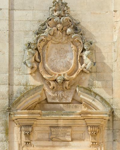 A close-up photograph of an ornate Baroque stone coat of arms and doorway lintel on a light-colored limestone building, featuring sculpted cherubs and a Latin inscription plaque reading "MARTINUS A MORBO BOVINO...".