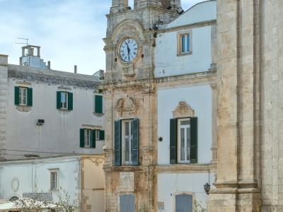 The image shows a sunlit Italian piazza with a prominent clock tower building featuring green shutters, people sitting on benches in the foreground, and a large stone column or wall on the right side.