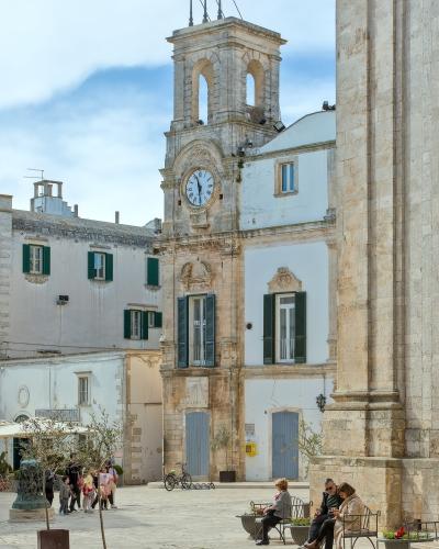 The image shows a sunlit Italian piazza with a prominent clock tower building featuring green shutters, people sitting on benches in the foreground, and a large stone column or wall on the right side.