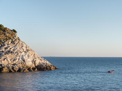 A rocky, sunlit coastal cliff with sparse vegetation on the left, overlooking a calm blue sea with a small red boat in the distance and a larger ship on the horizon under a clear sky.
