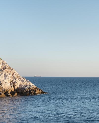A rocky, sunlit coastal cliff with sparse vegetation on the left, overlooking a calm blue sea with a small red boat in the distance and a larger ship on the horizon under a clear sky.
