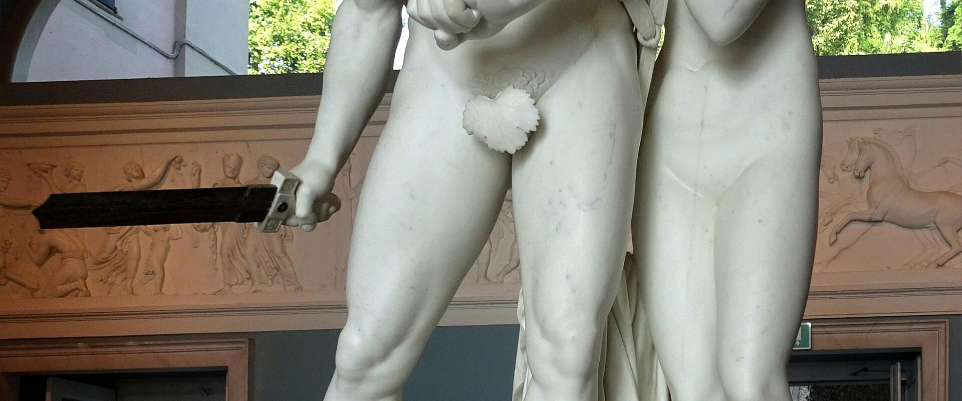 A white marble sculpture of Mars (Ares) and Venus (Aphrodite) standing together indoors, with the inscription "MARTE E VENERE" on the base, set against a decorated wall with arched windows showing greenery outside.