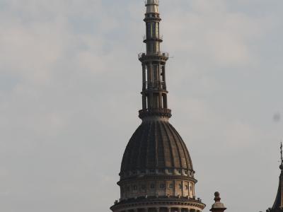 The image shows the distinctive dome and lantern of the Basilica di San Gaudenzio in Novara, Italy, featuring a large dark dome, a classical drum with columns, and a tall, ornate spire topped by a golden statue against a cloudy sky.