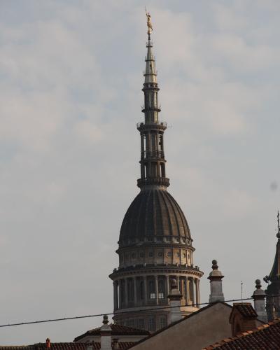 The image shows the distinctive dome and lantern of the Basilica di San Gaudenzio in Novara, Italy, featuring a large dark dome, a classical drum with columns, and a tall, ornate spire topped by a golden statue against a cloudy sky.