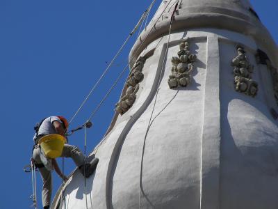 A worker in safety gear is rappelling down the white, bulbous dome of a Baroque-style church tower against a clear blue sky.