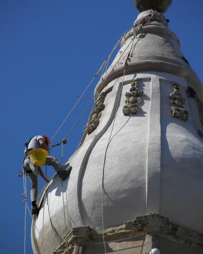 A worker in safety gear is rappelling down the white, bulbous dome of a Baroque-style church tower against a clear blue sky.