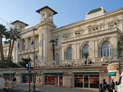 The image shows the exterior of a grand, ornate building with large signage reading "CASINO SANREMO" on the facade, palm trees, flags (including Italian and EU), and street-level activity under a clear sky.