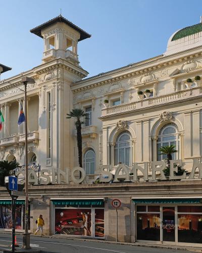 The image shows the exterior of a grand, ornate building with large signage reading "CASINO SANREMO" on the facade, palm trees, flags (including Italian and EU), and street-level activity under a clear sky.