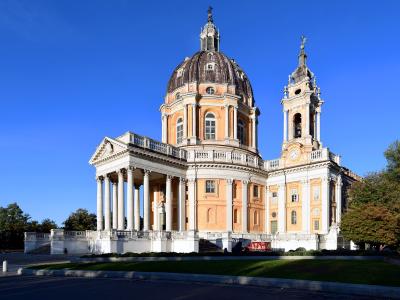 The image shows a large Baroque-style basilica with a prominent central dome, a tall bell tower, and a classical portico with columns, set against a clear blue sky with green lawns and trees in the foreground.