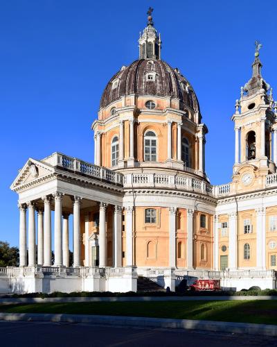 The image shows a large Baroque-style basilica with a prominent central dome, a tall bell tower, and a classical portico with columns, set against a clear blue sky with green lawns and trees in the foreground.