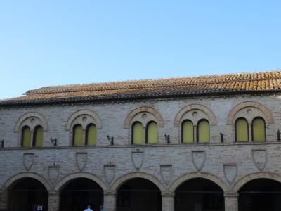 The image shows a two-story brick building with a loggia of arches on the ground floor and a row of arched windows on the upper floor, featuring decorative shields and small statues along the parapet.