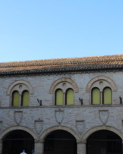 The image shows a two-story brick building with a loggia of arches on the ground floor and a row of arched windows on the upper floor, featuring decorative shields and small statues along the parapet.