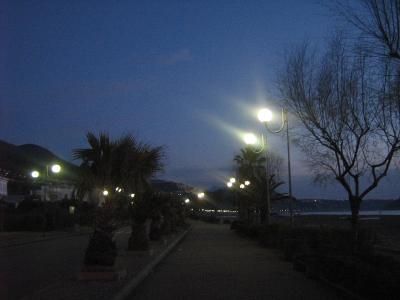 A paved coastal promenade at dusk, lined with palm trees and distinctive curved streetlights, with mountains in the background and the sea to the right.