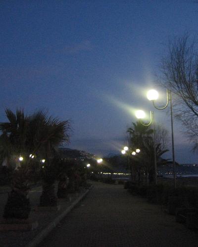 A paved coastal promenade at dusk, lined with palm trees and distinctive curved streetlights, with mountains in the background and the sea to the right.