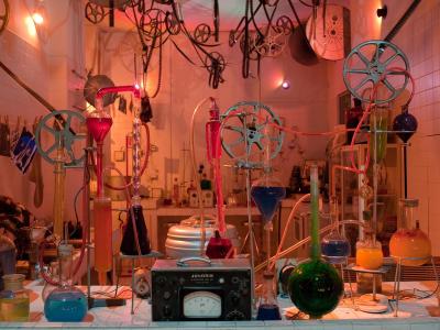 A laboratory-style art installation featuring glass beakers with colored liquids, film reels, and hanging film strips under red lighting, with a "JONGBIS" device in the foreground.
