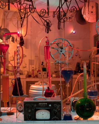 A laboratory-style art installation featuring glass beakers with colored liquids, film reels, and hanging film strips under red lighting, with a "JONGBIS" device in the foreground.