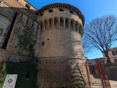 A low-angle shot of a large, cylindrical brick tower with corbelled battlements and a tiled roof, part of a larger fortress complex, with a sign in the foreground reading "ENTRATA PER VISITA ALLA ROCCA" (Entrance for visit to the fortress) and a r