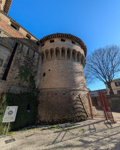 A low-angle shot of a large, cylindrical brick tower with corbelled battlements and a tiled roof, part of a larger fortress complex, with a sign in the foreground reading "ENTRATA PER VISITA ALLA ROCCA" (Entrance for visit to the fortress) and a r