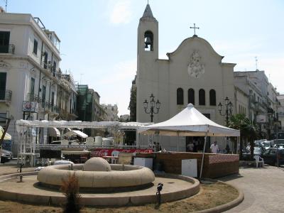 The image shows a sunlit public square with a distinctive circular stone fountain in the foreground, white event tents, and a prominent white church with a bell tower and a cross on its facade; signage for "Coca-Cola" and a "Conad" supermarket are