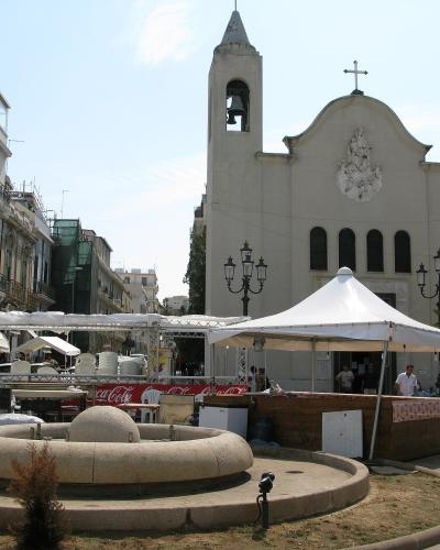 The image shows a sunlit public square with a distinctive circular stone fountain in the foreground, white event tents, and a prominent white church with a bell tower and a cross on its facade; signage for "Coca-Cola" and a "Conad" supermarket are
