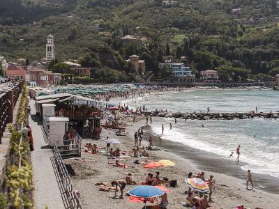 A crowded sandy beach with numerous sunbathers and umbrellas, backed by a town with a prominent bell tower and green hills, viewed from an elevated promenade.