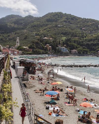 A crowded sandy beach with numerous sunbathers and umbrellas, backed by a town with a prominent bell tower and green hills, viewed from an elevated promenade.