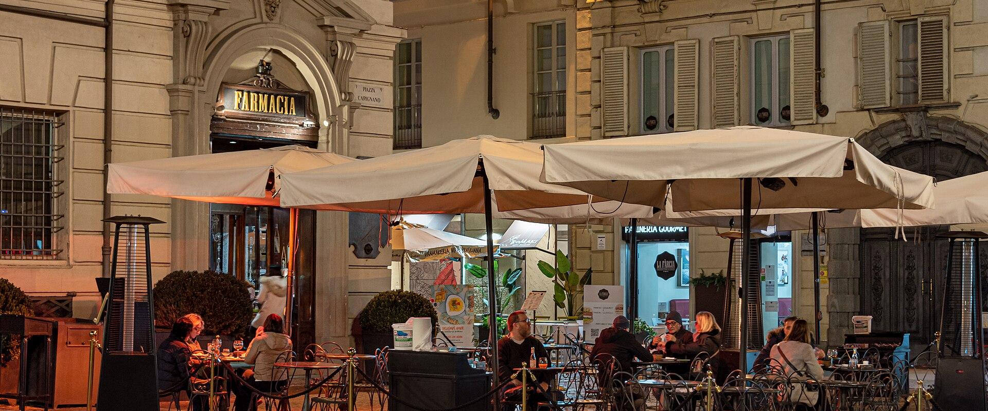 A nighttime view of an outdoor café seating area under large white umbrellas, with patrons dining at tables on a cobblestone plaza; visible signage includes "FARMACIA" above an arched doorway and a street sign reading "VIA CARIGNANO".