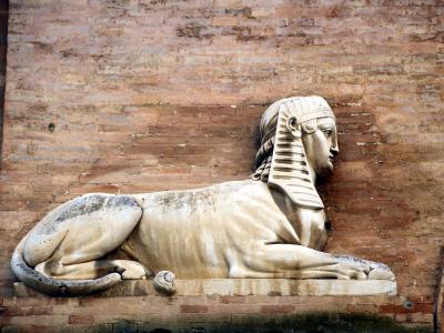 A stone sphinx sculpture with a human head and lion's body, mounted against a weathered brick wall.