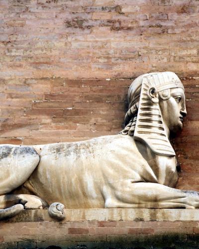 A stone sphinx sculpture with a human head and lion's body, mounted against a weathered brick wall.
