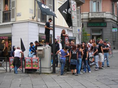A street scene in an Italian town showing a crowd gathered on a paved corner near buildings with signage including "CORSO DANTE," "SAN PAOLO," and "notivi," with people holding black flags and posters for an event.