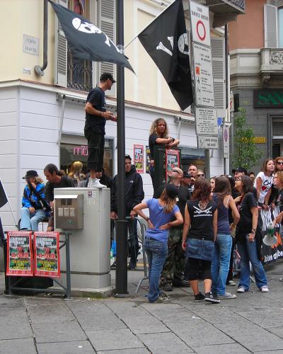 A street scene in an Italian town showing a crowd gathered on a paved corner near buildings with signage including "CORSO DANTE," "SAN PAOLO," and "notivi," with people holding black flags and posters for an event.