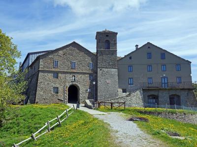 A stone church with a bell tower and adjacent building set on a grassy hillside under a blue sky, with a gravel path leading to the entrance.