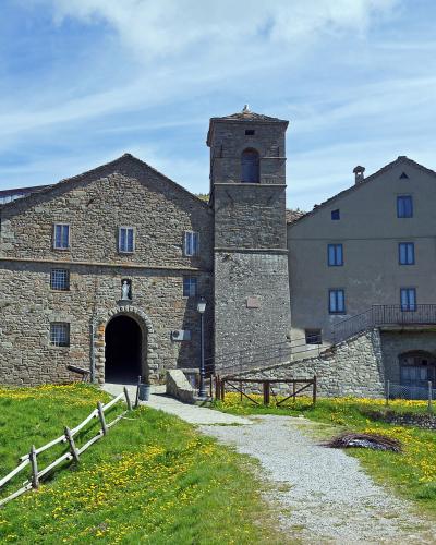 A stone church with a bell tower and adjacent building set on a grassy hillside under a blue sky, with a gravel path leading to the entrance.