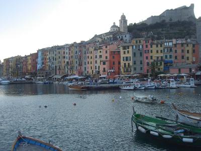 A harbor scene featuring a row of colorful multi-story buildings along the waterfront, with boats moored in the foreground and a hilltop fortress and church visible in the background.