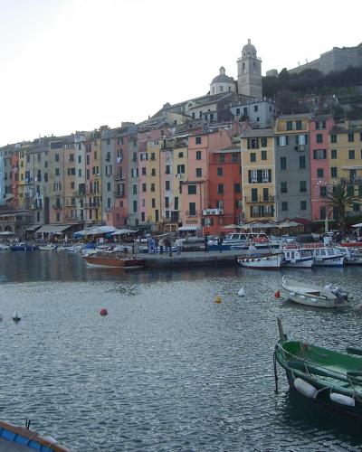 A harbor scene featuring a row of colorful multi-story buildings along the waterfront, with boats moored in the foreground and a hilltop fortress and church visible in the background.