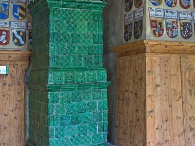 A tall, green-glazed ceramic tiled stove (Kachelofen) stands in a room with wooden paneling and walls adorned with painted coats of arms bearing Germanic names like "Brixen," "Freiegg," and "De Pauli."
