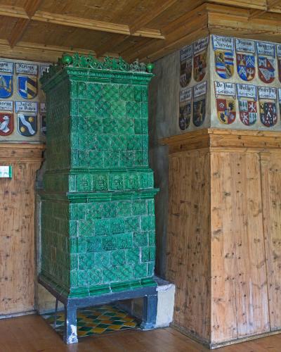 A tall, green-glazed ceramic tiled stove (Kachelofen) stands in a room with wooden paneling and walls adorned with painted coats of arms bearing Germanic names like "Brixen," "Freiegg," and "De Pauli."