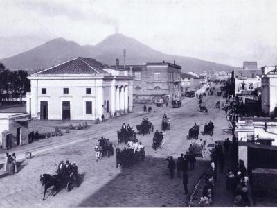 A black-and-white historical photograph showing a wide cobblestone street with horse-drawn carriages and pedestrians, dominated by a large neoclassical building with columns on the left, set against the backdrop of Mount Vesuvius.