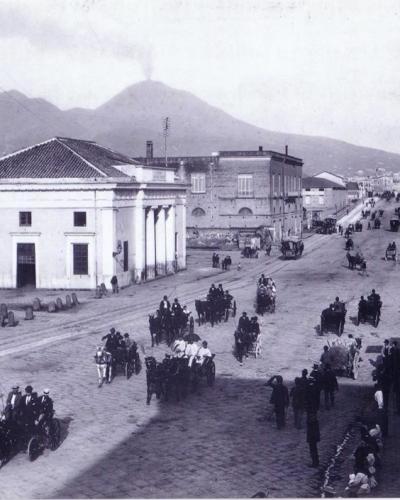 A black-and-white historical photograph showing a wide cobblestone street with horse-drawn carriages and pedestrians, dominated by a large neoclassical building with columns on the left, set against the backdrop of Mount Vesuvius.