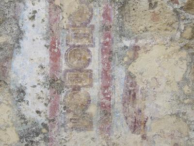 A close-up photograph of a weathered, peeling wall surface showing remnants of faded Roman-style frescoes with geometric patterns and faint figures in red, ochre, and blue pigments.