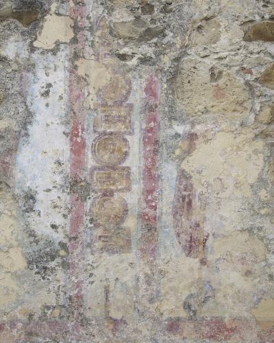 A close-up photograph of a weathered, peeling wall surface showing remnants of faded Roman-style frescoes with geometric patterns and faint figures in red, ochre, and blue pigments.