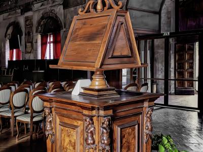 The image shows a large, ornate wooden lectern (reading desk) with carved figures, situated indoors on a protective plastic sheet, with rows of chairs and arched windows in the background.
