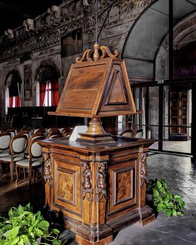 The image shows a large, ornate wooden lectern (reading desk) with carved figures, situated indoors on a protective plastic sheet, with rows of chairs and arched windows in the background.