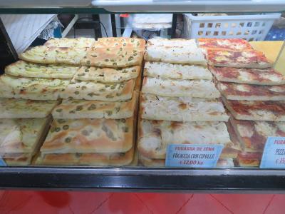 A display case in a bakery or food shop showing stacks of various rectangular flatbreads, including one labeled "FUGÀSSA DE ZÉNA" (Focaccia with onions) priced at €12.00/kg.